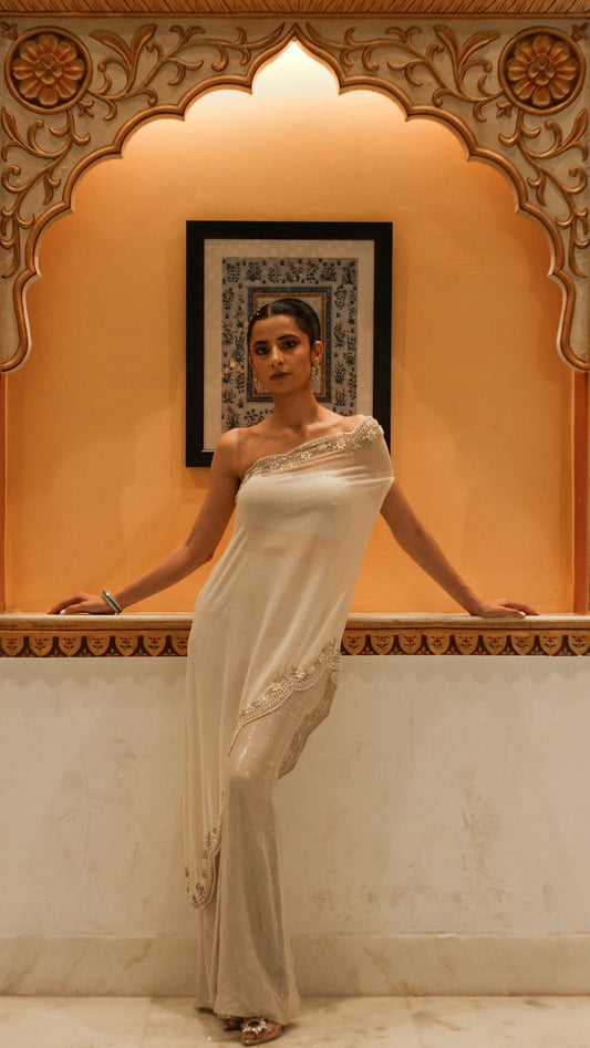 Woman in a white saree standing in front of an ornate wall design.
