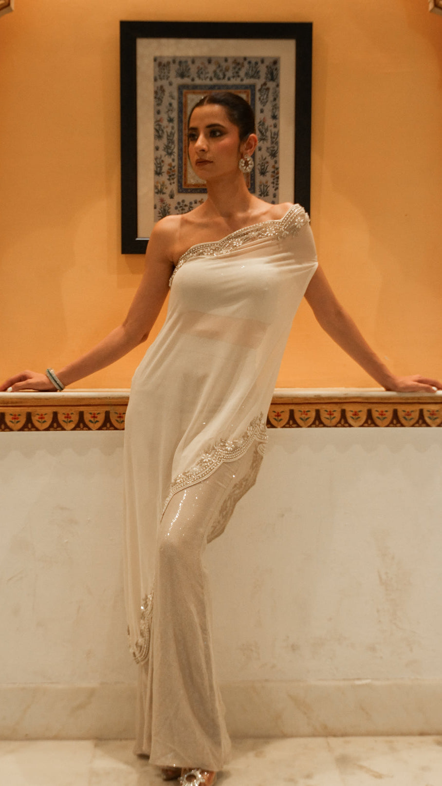 Woman in a white saree with intricate designs standing in an ornate room.