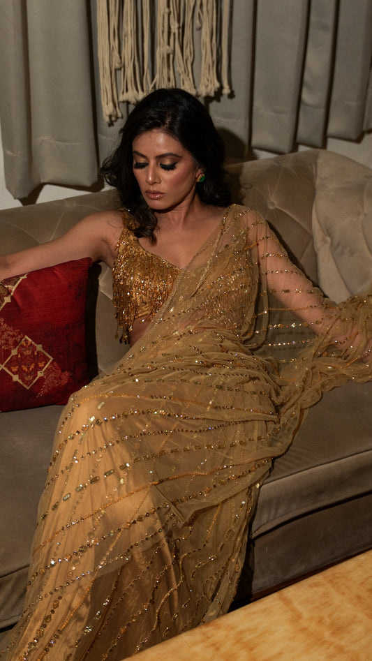 Woman in a gold saree sitting on a couch in a room with curtains and a pillow.