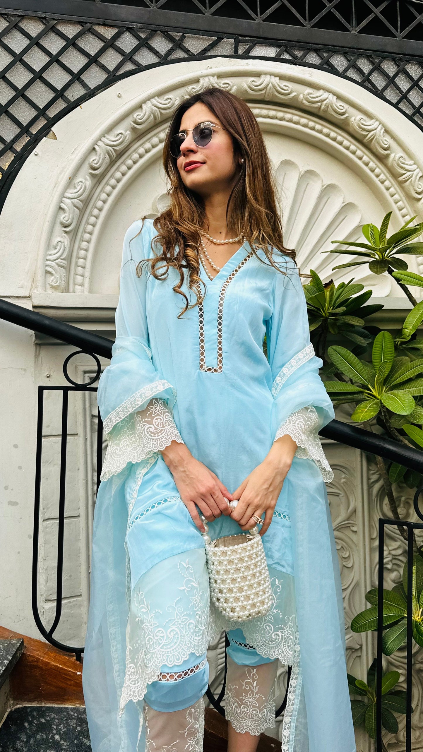 Woman in light blue outfit standing in front of decorative archway with plants.
