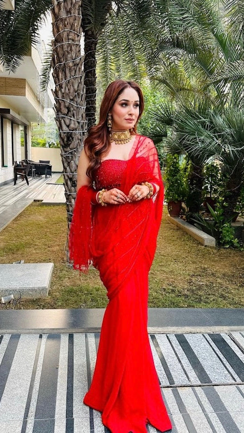 Woman in a red saree standing outdoors with palm trees and a building in the background