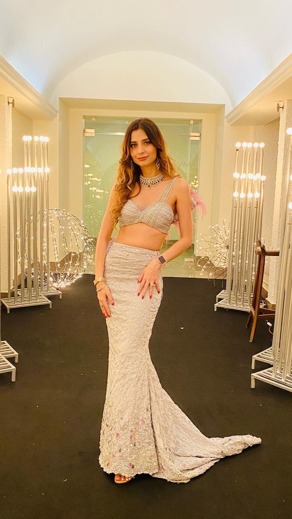 Woman in a silver dress standing in an elegant interior setting with decorative lights.