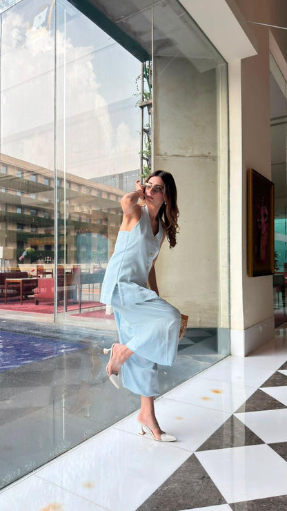 A woman in a light blue sleeveless dress and white heels poses dynamically on a black-and-white checkered floor next to a large glass window in a modern building.